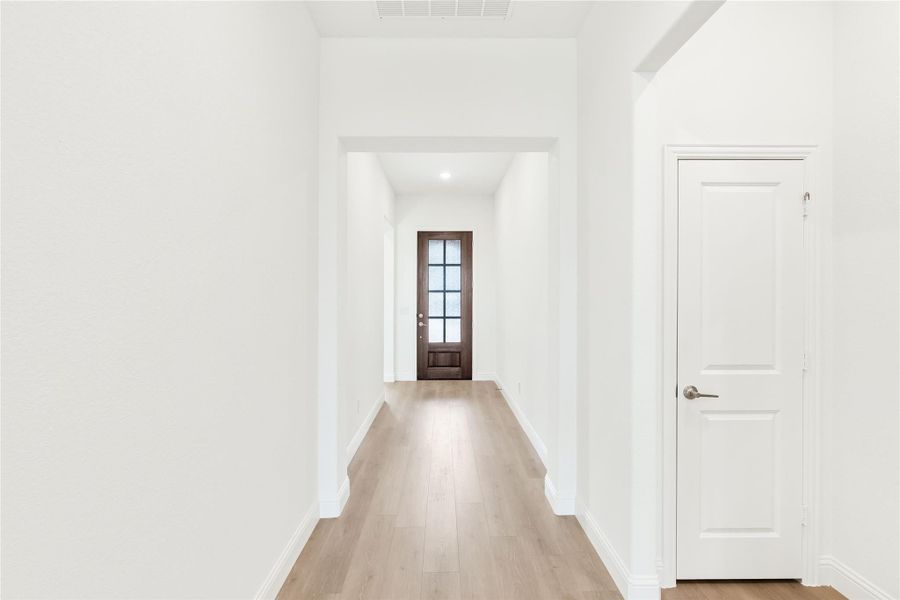 Hallway with white walls, light wood floors, and dark wood front door with glass panels at the end Hallway with white walls, light wood floors, and dark wood front door with glass panels at the end