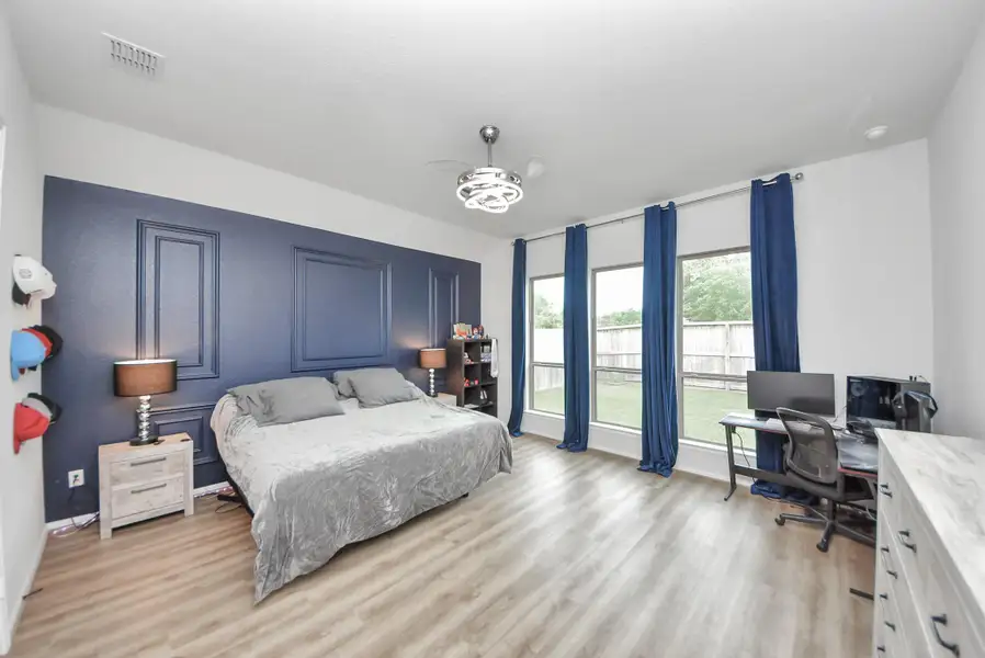 This spacious bedroom features a stylish custom dark accent wall, wood flooring, and generous natural light from large windows with blue curtains. It includes a comfortable bed, modern ceiling light, and a desk area perfect for work or study.