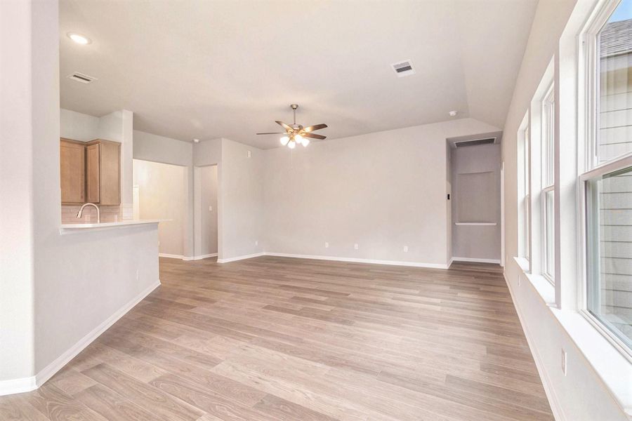 This photo shows a bright, open-concept living space with wood flooring, neutral walls, and a ceiling fan. It features a modern kitchen with a breakfast bar and large windows for natural light.