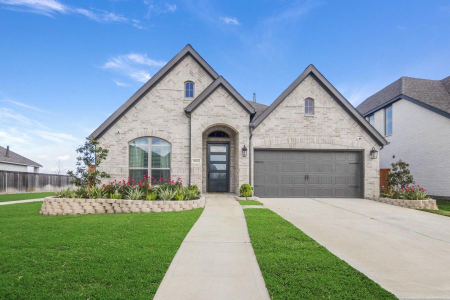 This is a modern, single-story home with a light brick exterior and dark gray gabled roof. It features a welcoming front entrance with a large window, a two-car garage, and a well-maintained front yard with landscaped flowerbeds.
