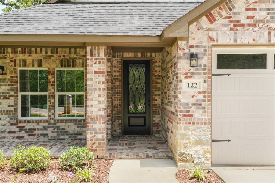 Brick exterior featuring a covered porch with a patterned brick floor, a dark-paneled entry door with decorative glass, two double-hung windows, and an attached garage with a window panel and decorative hardware Brick exterior featuring a covered porch with a patterned brick floor, a dark-paneled entry door with decorative glass, two double-hung windows, and an attached garage with a window panel and decorative hardware