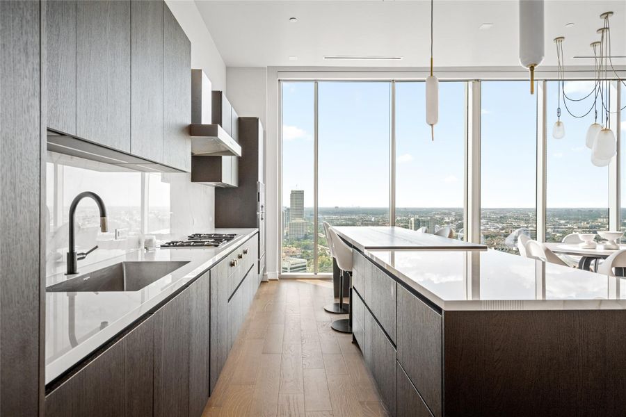 This modern high-rise kitchen exudes sophistication with warm brown cabinetry and elegant porcelain countertops, featuring Gaggenau appliances, a gas cooktop, and induction range. It flows seamlessly into the dining area and is illuminated by natural light from floor-to-ceiling windows that frame expansive city views.