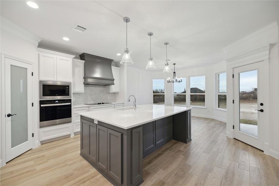 Kitchen featuring a center island with sink, white cabinetry, appliances with stainless steel finishes, custom range hood, and pendant lighting