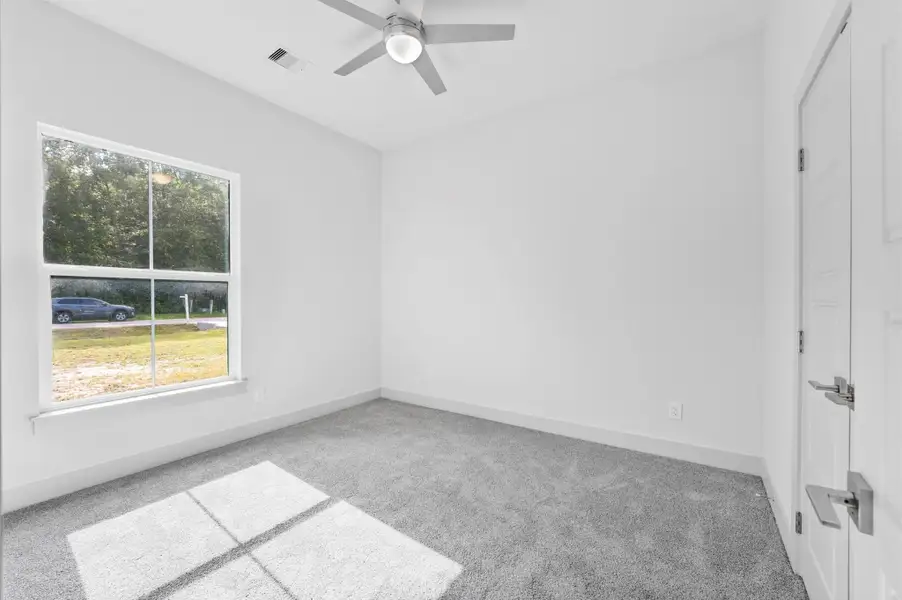 This is a bright, empty room with light gray carpeting, neutral walls, a large window providing natural light, and a ceiling fan. Ideal for a bedroom or office space. This is a bright, empty room with light gray carpeting, neutral walls, a large window providing natural light, and a ceiling fan. Ideal for a bedroom or office space.