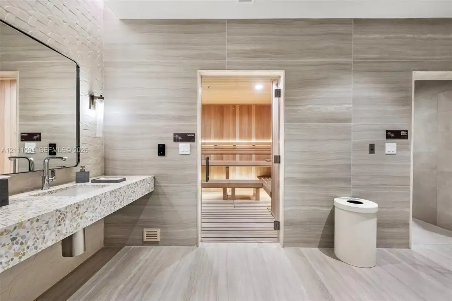 Sauna room featuring cedar wood wall paneling and tiered bench seating, complemented by a stone-tiled accent wall, a floating vanity with a terrazzo-style countertop, and a large wall-mounted mirror Sauna room featuring cedar wood wall paneling and tiered bench seating, complemented by a stone-tiled accent wall, a floating vanity with a terrazzo-style countertop, and a large wall-mounted mirror