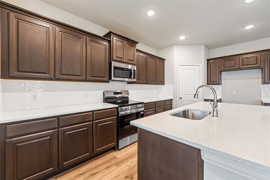 Kitchen featuring appliances with stainless steel finishes, dark brown cabinetry, light stone counters, light wood-style floors, and recessed lighting