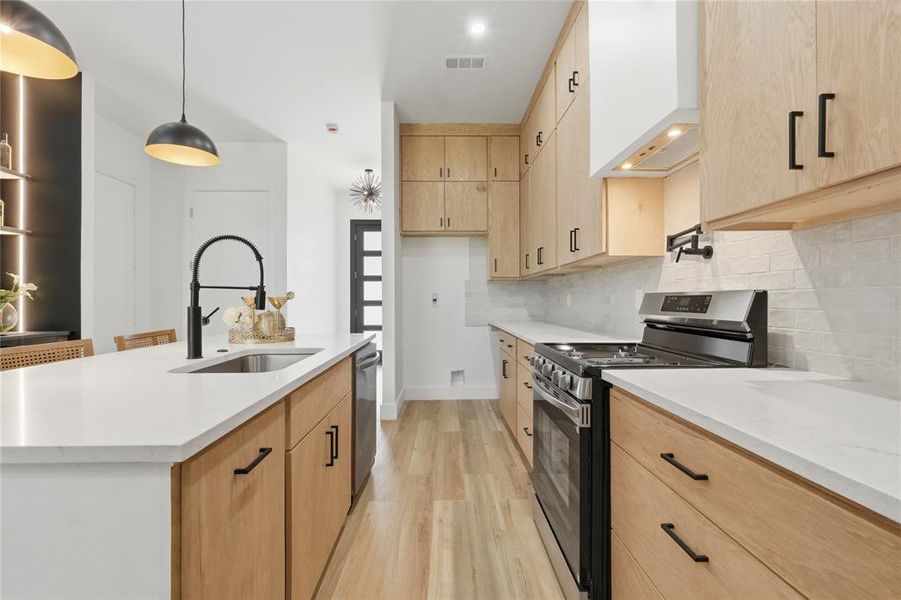 Modern kitchen featuring a large island with an undermount sink and matte black faucet, light wood-finish cabinetry, white countertops, and a stainless steel oven range