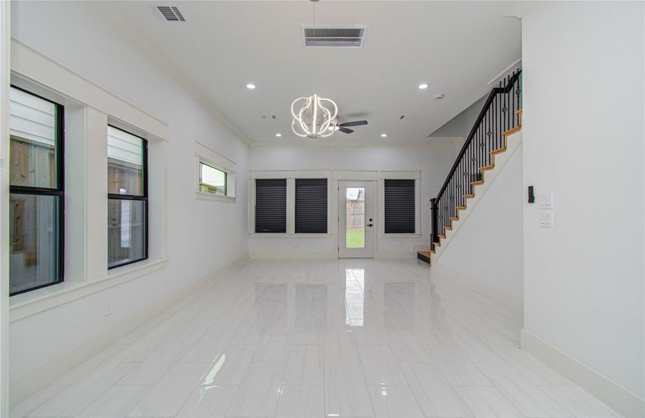This bright and spacious living area features sleek white tile flooring, a modern light fixture, and large windows with dark blinds. The room opens to a backyard through glass doors and includes a staircase with elegant iron railings, adding a touch of sophistication.