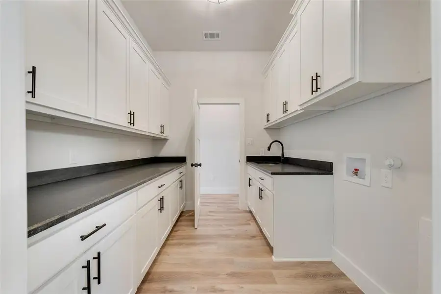 Spacious utility room featuring extensive white cabinetry with matte black hardware, dark countertops, and wood-finish flooring