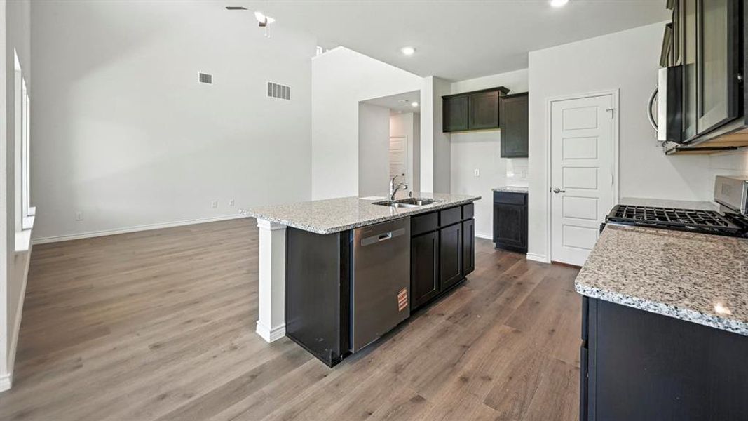 Open-concept kitchen and living area featuring wood-finish flooring, a kitchen island with integrated sink and dishwasher, granite countertops, dark cabinetry, and a gas range