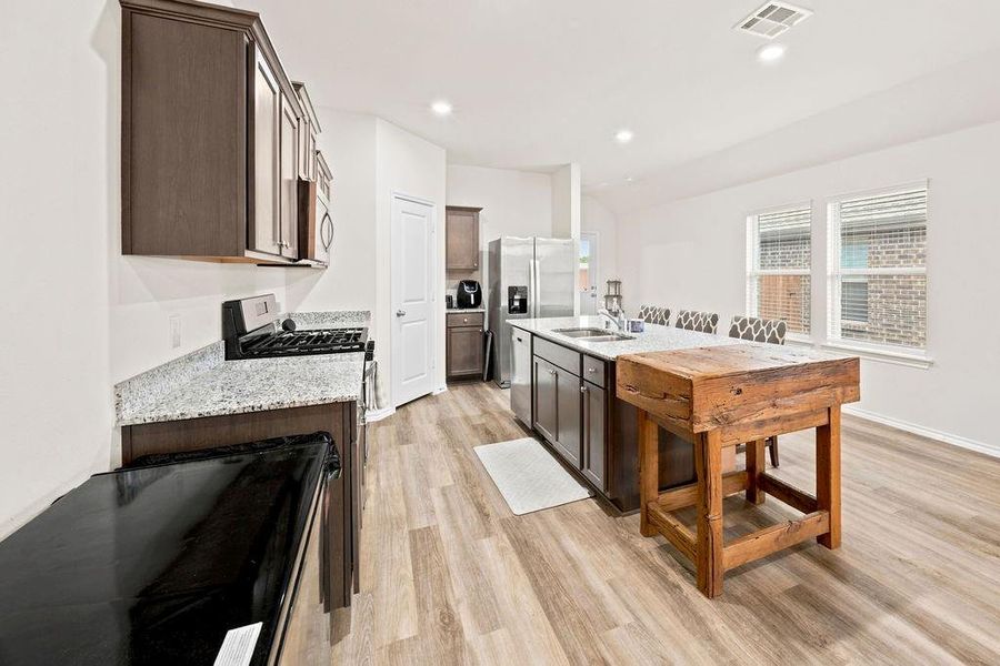 Open-concept kitchen featuring dark wood cabinetry, light granite countertops, stainless steel appliances, and wood-finish flooring Open-concept kitchen featuring dark wood cabinetry, light granite countertops, stainless steel appliances, and wood-finish flooring