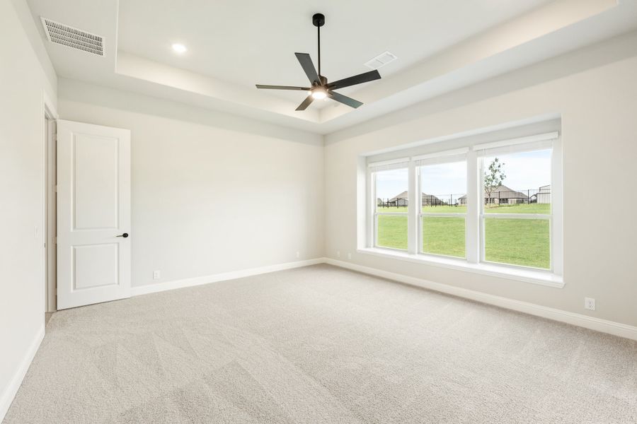 Empty bedroom with tray ceiling, ceiling fan, carpet flooring, and large window overlooking green yard Empty bedroom with tray ceiling, ceiling fan, carpet flooring, and large window overlooking green yard