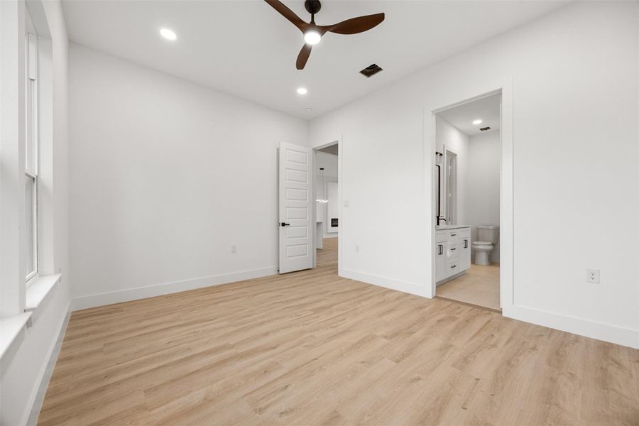 Bright, modern bedroom with light wood-like flooring and white walls. It features a ceiling fan, large windows, and an en-suite bathroom with a contemporary vanity.