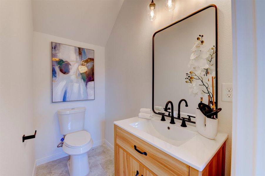 First-floor powder bath with wood-tone vanity, white quartz countertop, matte black faucet, large black-framed mirror, pendant lighting, and vaulted ceiling detail.