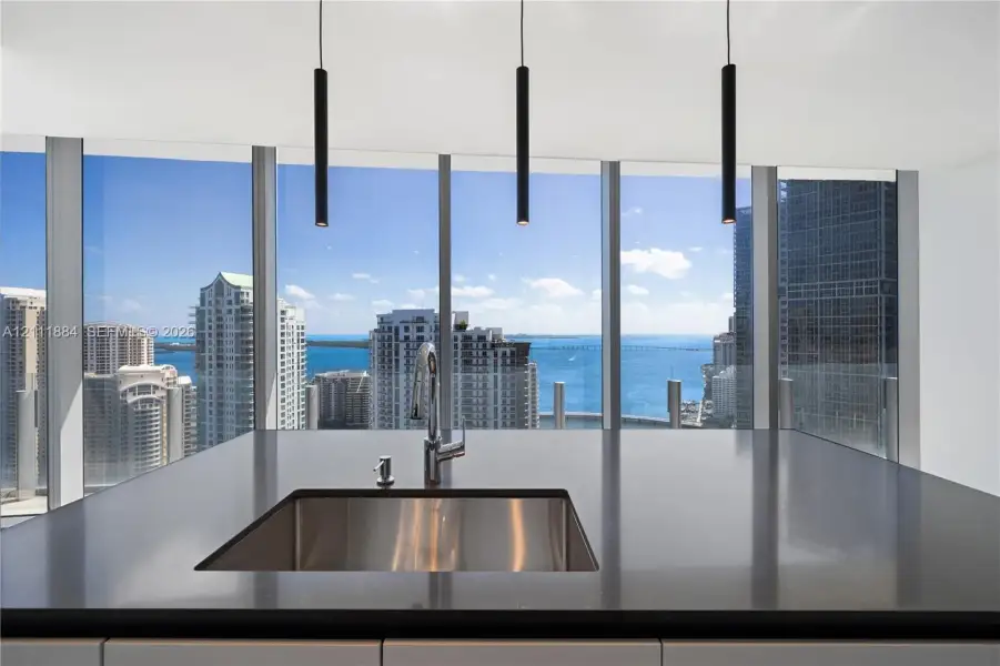 Kitchen island featuring dark solid-surface countertops, an undermount stainless steel sink with a high-arch faucet, floor-to-ceiling windows, and matte black cylindrical pendant lighting Kitchen island featuring dark solid-surface countertops, an undermount stainless steel sink with a high-arch faucet, floor-to-ceiling windows, and matte black cylindrical pendant lighting