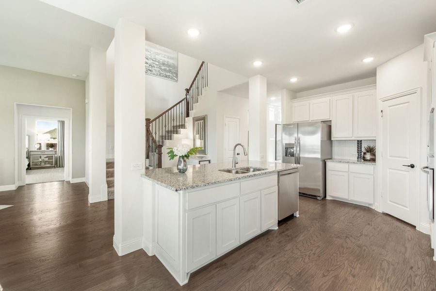 Kitchen with white cabinets, granite island, stainless steel appliances, and dark hardwood floors open to staircase.