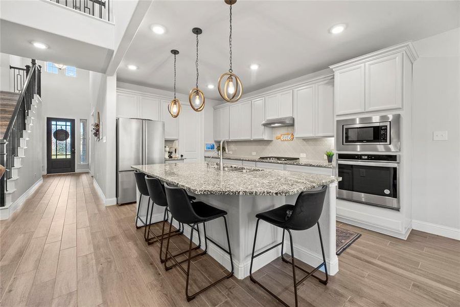 This is a modern, spacious kitchen featuing, stainless steel appliances, and an island with seating. The room is well-lit with natural light from the entrance and stylish pendant lights. There's an open floor plan with a view of the staircase and upper level, offering a sense of openness and connectivity.