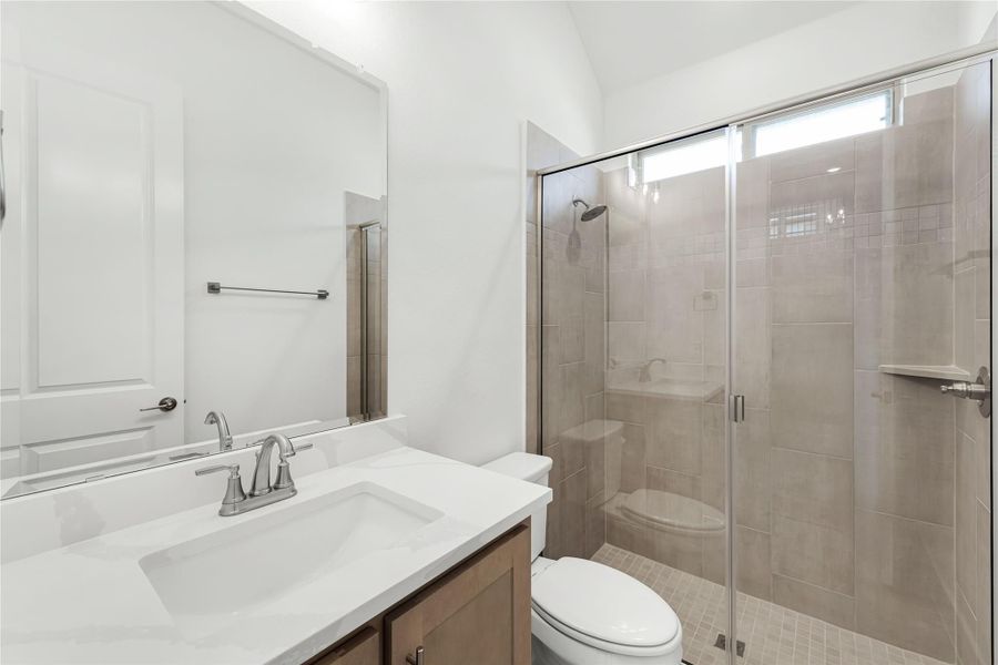 Bathroom with white quartz vanity, wood cabinets, toilet, and glass-enclosed tile shower with vaulted ceiling