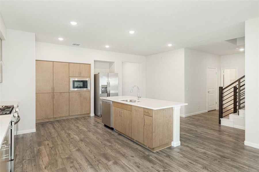 Open kitchen with light wood cabinets, center island with sink, stainless steel appliances, and hardwood floors