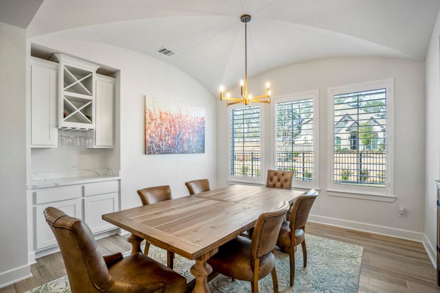 This bright dining room features a wooden table with six cushioned chairs, large windows with plantation shutters, a modern light fixture, and built-in cabinetry with a wine rack, creating a cozy and functional space.
