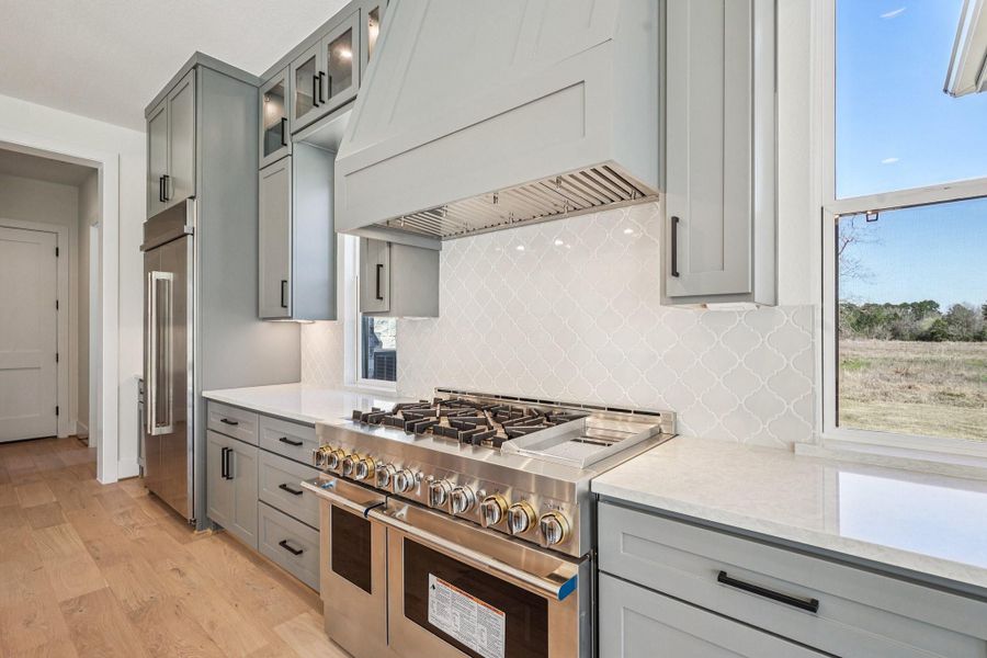 This modern kitchen features sleek gray cabinetry, a high-end stainless steel gas range, and light countertops. The elegant tile backsplash adds a touch of sophistication, and large windows provide ample natural light with scenic views.
