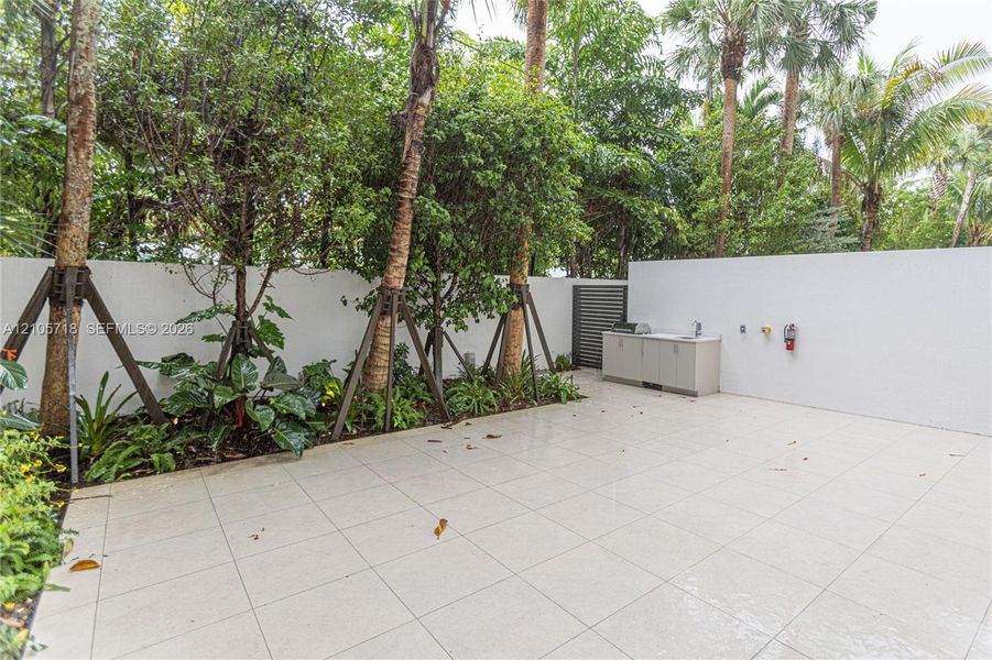 Private outdoor patio featuring light-toned tile flooring, built-in cabinetry with a sink, stainless steel grill, solid privacy walls, and mature perimeter landscaping