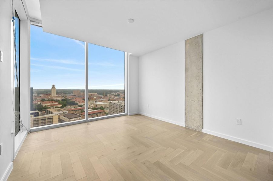 Step into this sleek and contemporary bedroom featuring floor-to-ceiling windows that frame spectacular views of the city and beyond. The natural light floods the room, enhancing the airy ambiance created by the light wood flooring and neutral color palette. The exposed concrete accent wall adds an industrial touch to the minimalist design.