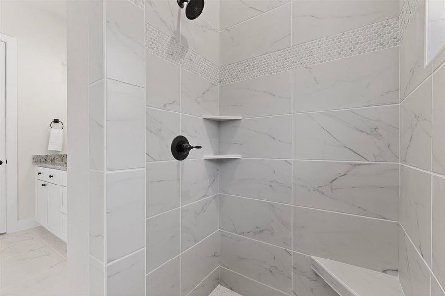 Spacious walk-in shower featuring large format tile with subtle veining, decorative mosaic accent band, built-in corner shelving, integrated shower bench, and dark bronze fixtures