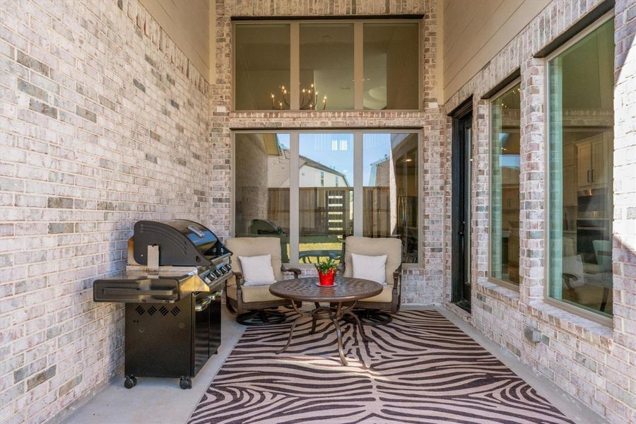 This cozy outdoor patio features brick walls, a grill, and comfortable seating around a small table on a decorative rug. Large windows and a glass door provide natural light and easy indoor access.