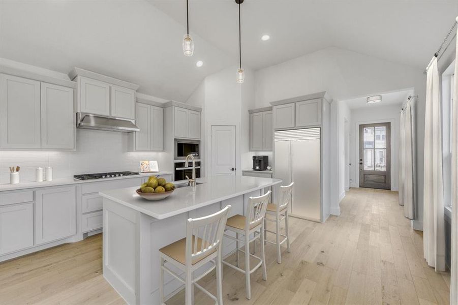 Virtually staged photo - Kitchen featuring a kitchen breakfast bar, stainless steel appliances, light wood-style floors, decorative light fixtures, and lofted ceiling