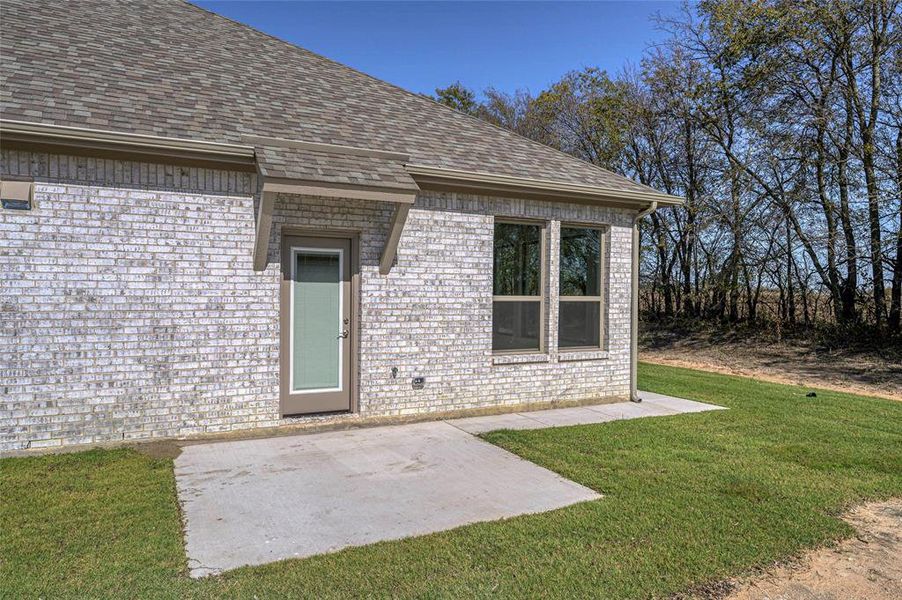 Entrance to property featuring a patio, a shingled roof, brick siding, and a yard Entrance to property featuring a patio, a shingled roof, brick siding, and a yard
