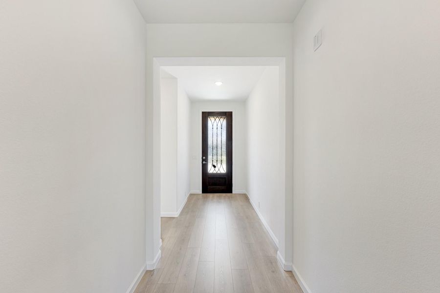 Hallway with light wood floors leading to a dark front door with decorative glass panel Hallway with light wood floors leading to a dark front door with decorative glass panel