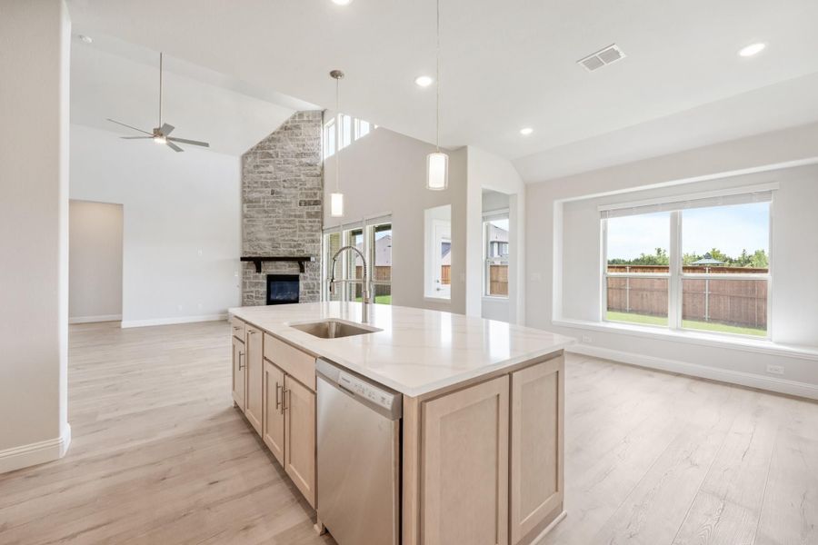 Kitchen island with sink and light wood cabinets, open to living area with stone fireplace and vaulted ceiling