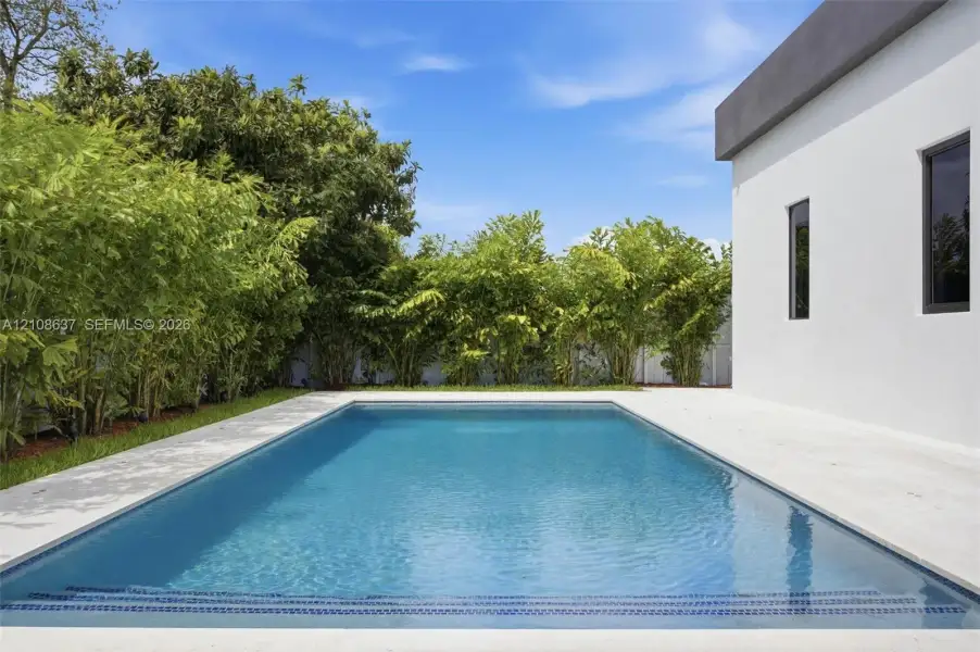 Rectangular in-ground pool featuring a stone-paver deck, crisp white exterior siding, and dense perimeter landscaping