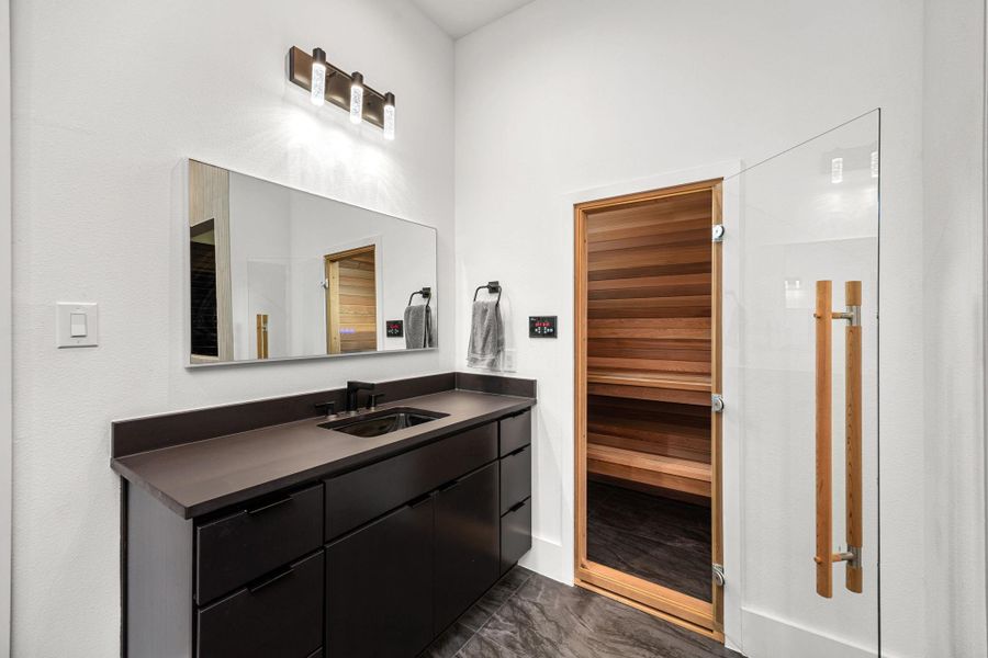 Modern bathroom with sleek dark cabinetry, large mirror, and elegant lighting. Features a glass door leading to a wood-paneled sauna, adding luxury and relaxation. Modern bathroom with sleek dark cabinetry, large mirror, and elegant lighting. Features a glass door leading to a wood-paneled sauna, adding luxury and relaxation.