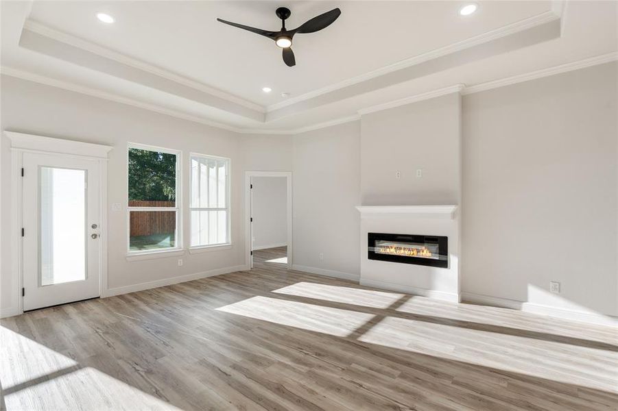 Living room featuring a tray ceiling, a glass covered fireplace, light wood-style floors, a ceiling fan, and crown molding