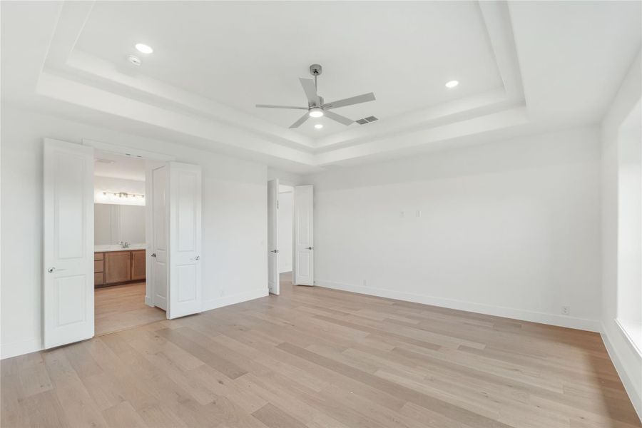 Empty bedroom with tray ceiling, ceiling fan, hardwood floors, and open door to ensuite bathroom