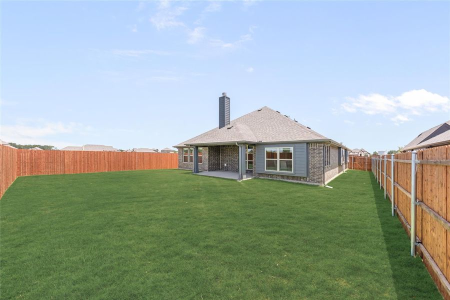 Backyard of brick home with large green lawn, covered patio, chimney, and wood privacy fence on both sides.