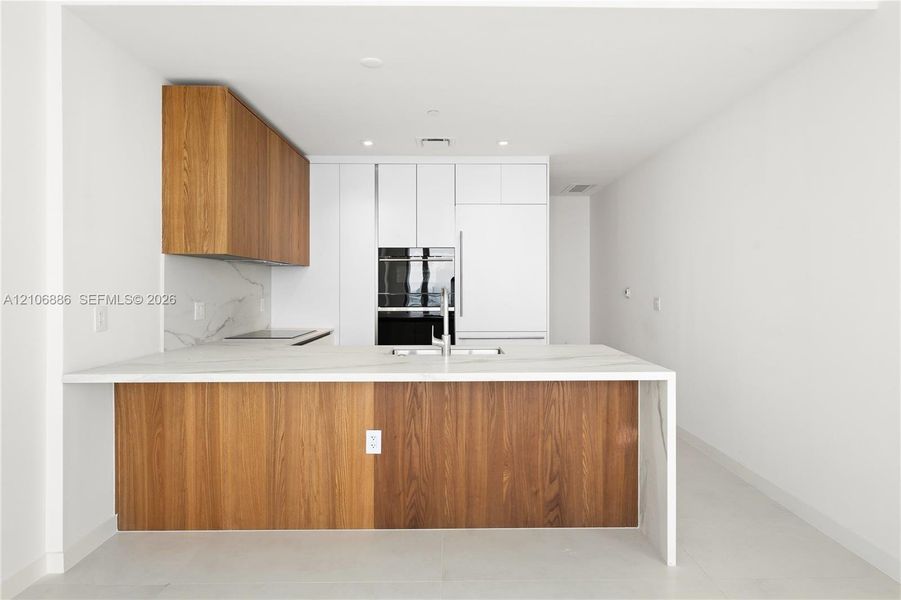 Kitchen featuring a center island with a stone-finish countertop and waterfall edge, two-tone wood-finish and flat-panel cabinetry, built-in wall oven, and integrated refrigerator