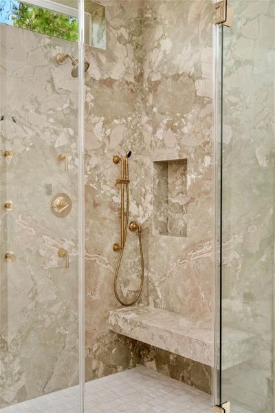 The primary shower makes a striking statement with richly veined natural stone enveloping the walls and dual floating benches. Waterworks brass fixtures, including dual shower heads, a hand-held shower, and body sprays, create an indulgent spa experience, while the frameless glass enclosure keeps the dramatic stonework at center stage. The primary shower makes a striking statement with richly veined natural stone enveloping the walls and dual floating benches. Waterworks brass fixtures, including dual shower heads, a hand-held shower, and body sprays, create an indulgent spa experience, while the frameless glass enclosure keeps the dramatic stonework at center stage.