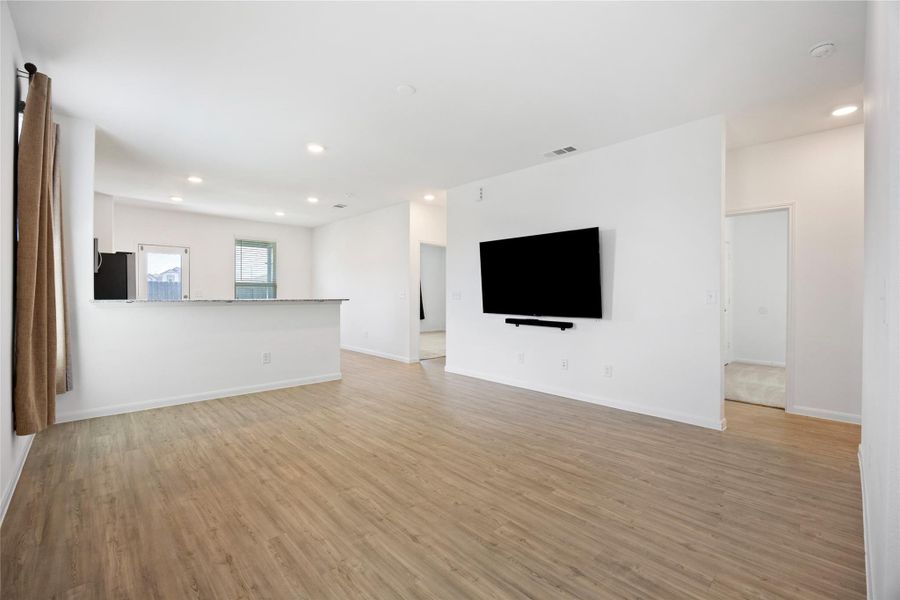 Bright and spacious open-concept living area with wood-style flooring, a mounted TV, and an adjacent kitchen featuring a breakfast bar. Large windows provide ample natural light.