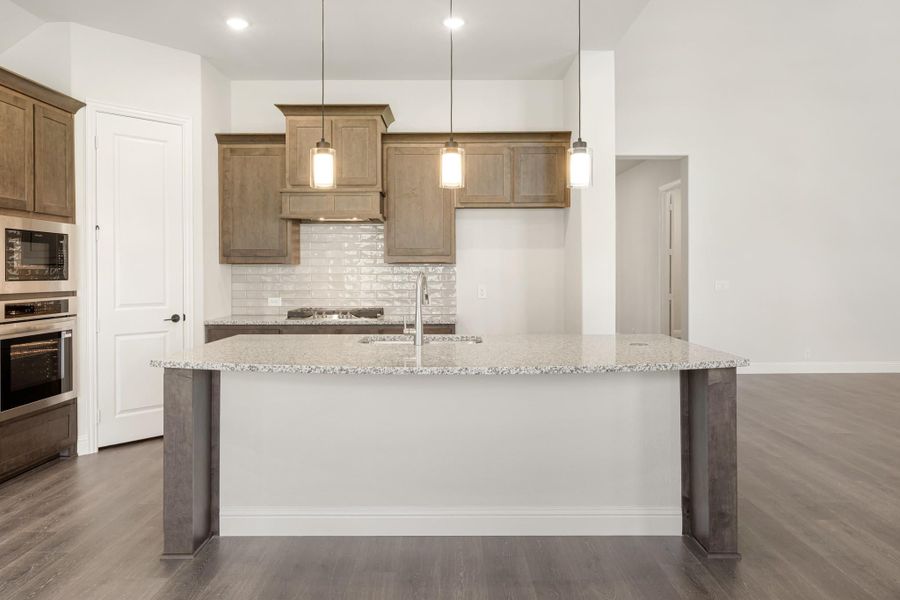 Kitchen with large granite island, brown wood cabinets, tile backsplash, and three pendant lights overhead Kitchen with large granite island, brown wood cabinets, tile backsplash, and three pendant lights overhead