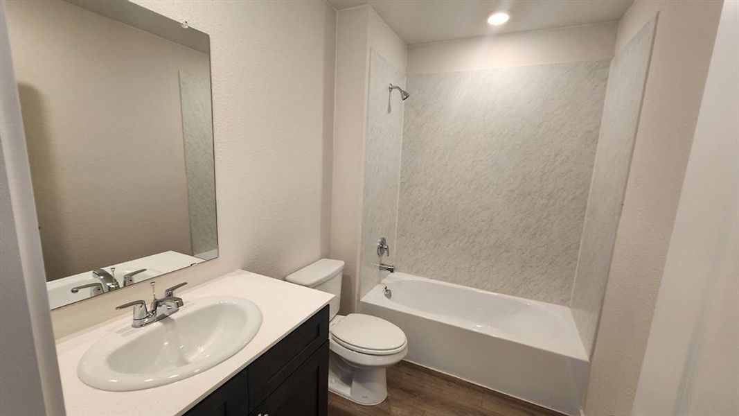 Bathroom featuring a white bathtub with a tile-patterned surround, a dark wood-finish vanity with a white countertop and integrated sink, a rectangular wall-mounted mirror, and wood-finish flooring Bathroom featuring a white bathtub with a tile-patterned surround, a dark wood-finish vanity with a white countertop and integrated sink, a rectangular wall-mounted mirror, and wood-finish flooring