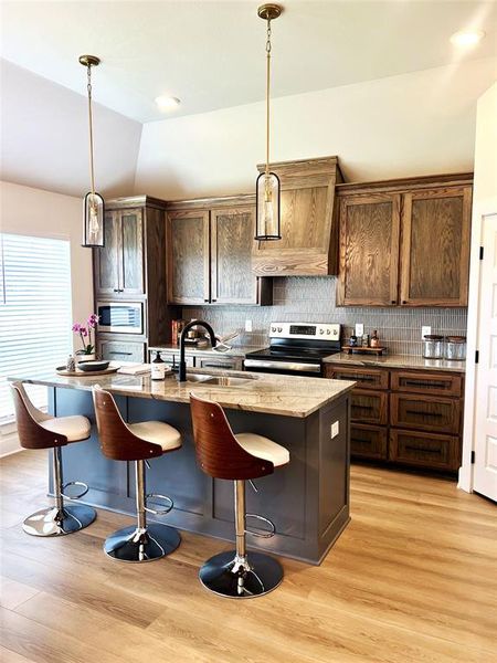 Kitchen featuring stainless steel appliances, a center island with sink, custom exhaust hood, light wood-type flooring, and lofted ceiling Kitchen featuring stainless steel appliances, a center island with sink, custom exhaust hood, light wood-type flooring, and lofted ceiling