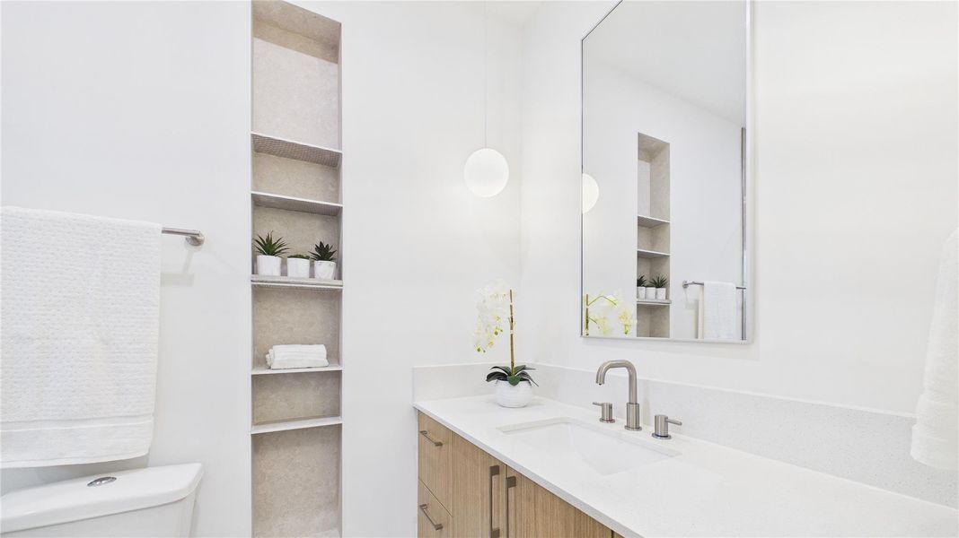 This is a modern bathroom featuring a sleek white countertop with a built-in sink, a large mirror, and minimalist shelving. The space is bright and airy, with a contemporary light fixture and subtle decorative touches.