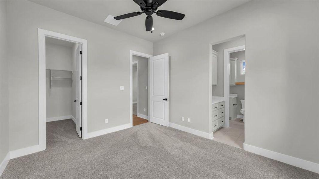 Carpeted room featuring a ceiling fan with light, an open doorway to a closet with wire shelving, and an open doorway revealing a bathroom with light-toned flooring, a vanity with drawers, and a toilet