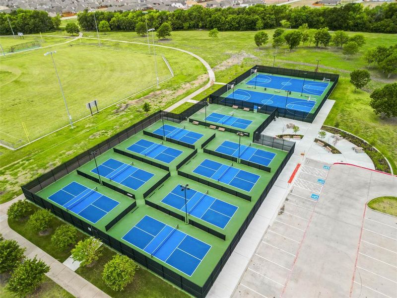 Recreational facility featuring multiple pickleball courts with blue playing surfaces and green surrounds, two basketball courts, an expansive grassy field, paved walking paths, and a dedicated parking area