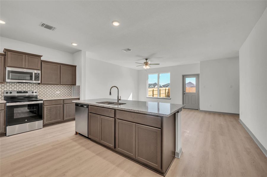 Kitchen featuring appliances with stainless steel finishes, an island with sink, backsplash, light stone countertops, and light wood-style flooring