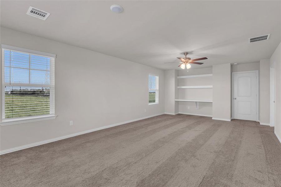 This is a spacious, well-lit  primary bedroom with neutral walls and carpet flooring. It features a large window for natural light, a ceiling fan for comfort, and built-in shelving for storage or decor.