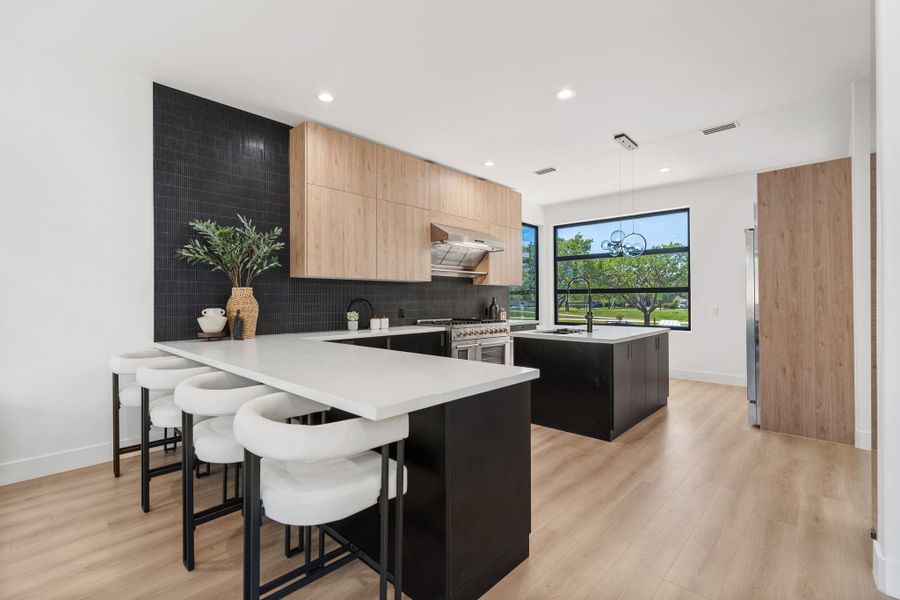 The kitchen's full perimeter reads clearly from here — black lower cabinetry, warm wood uppers, black vertical tile backsplash, and a large black-framed adjacent to the sink that pulls greenery directly into the workspace and bathes the kitchen in natural light.